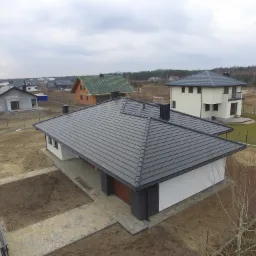 Nowoczesny dom jednorodzinny z grafitowym dachem z blachodachówki modułowej, widok z góry na działkę w stanie surowym i okoliczną zabudowę.