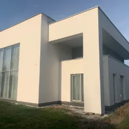 Nowoczesny, biały dom jednorodzinny z dużymi oknami, minimalistyczna bryła, widoczne świeżo wykonane prace elewacyjne, trawnik z resztkami materiałów budowlanych.