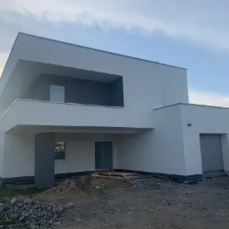 Nowoczesny, dwukondygnacyjny dom z płaskim dachem, świeżo po tynkowaniu elewacji na biało, z szarymi elementami wykończeniowymi, widoczne dwie bramy garażowe i niedokończone otoczenie.