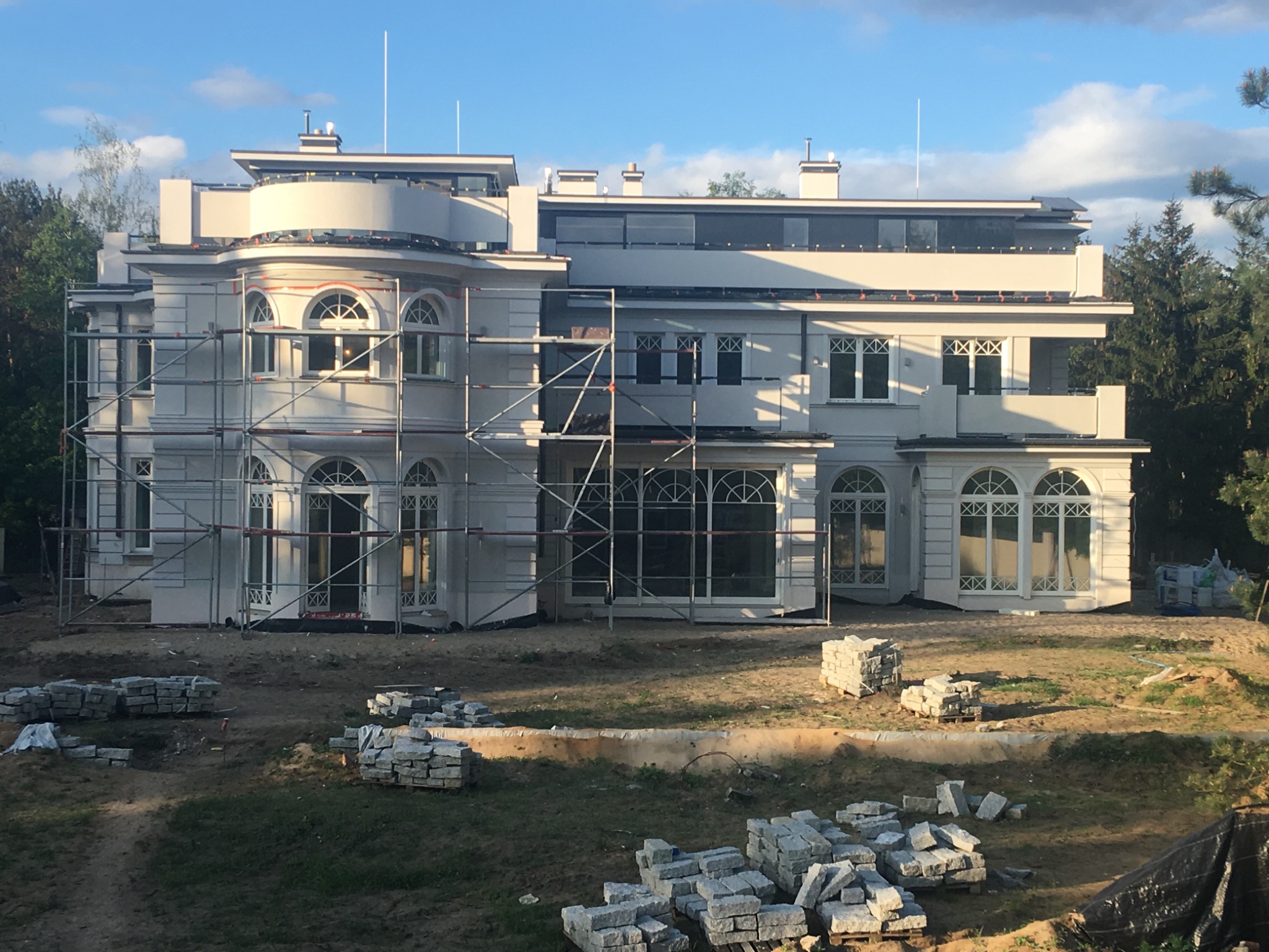 Elewacja w trakcie prac wykończeniowych: biały budynek z rusztowaniami, widoczne sterty kamieni na podwórku, słoneczny dzień.