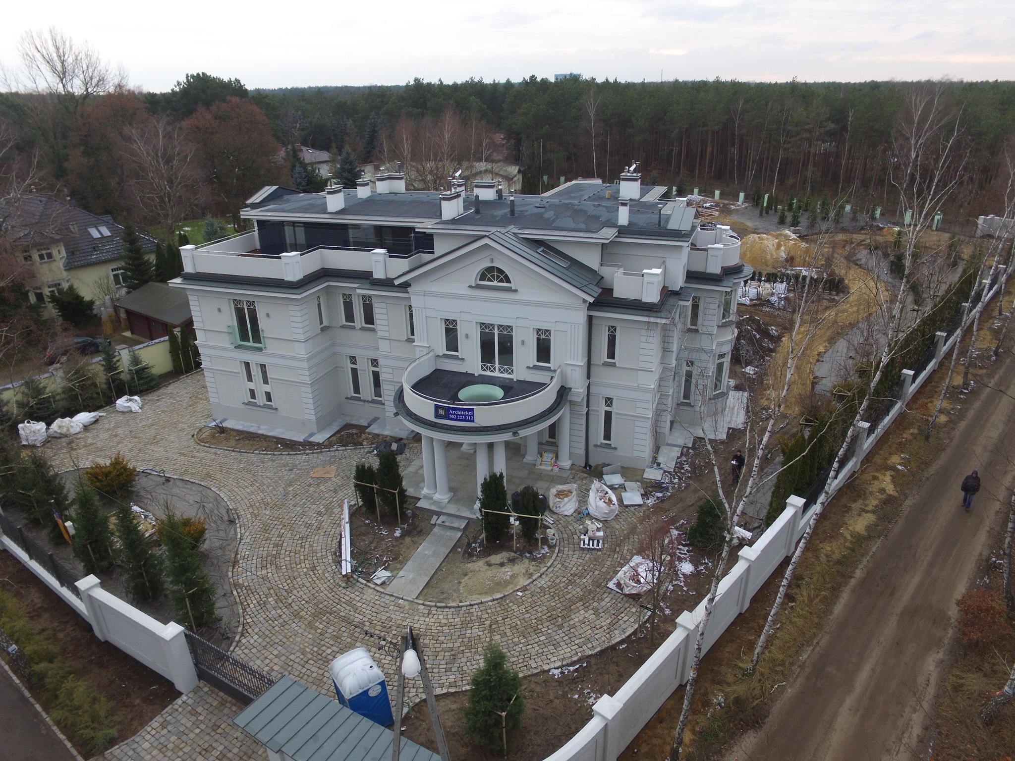 Widok z lotu ptaka na duży, dwupiętrowy dom w stylu klasycystycznym z płaskim dachem i balkonem z kolumnami, otoczony brukowanym podjazdem i ogrodzeniem, w trakcie prac wykończeniowych ogrodu.