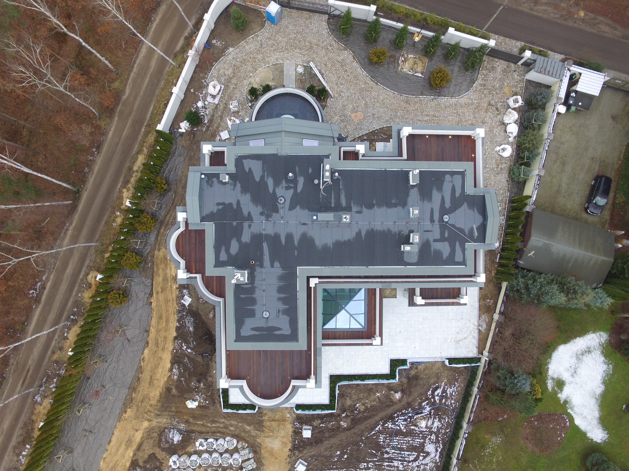 Widok z drona na rozległy, nowoczesny dom z płaskim, ciemnoszarym dachem, drewnianymi tarasami i szklanym świetlikiem, otoczony brukowanym podjazdem, uporządkowanym ogrodem i częściowo...