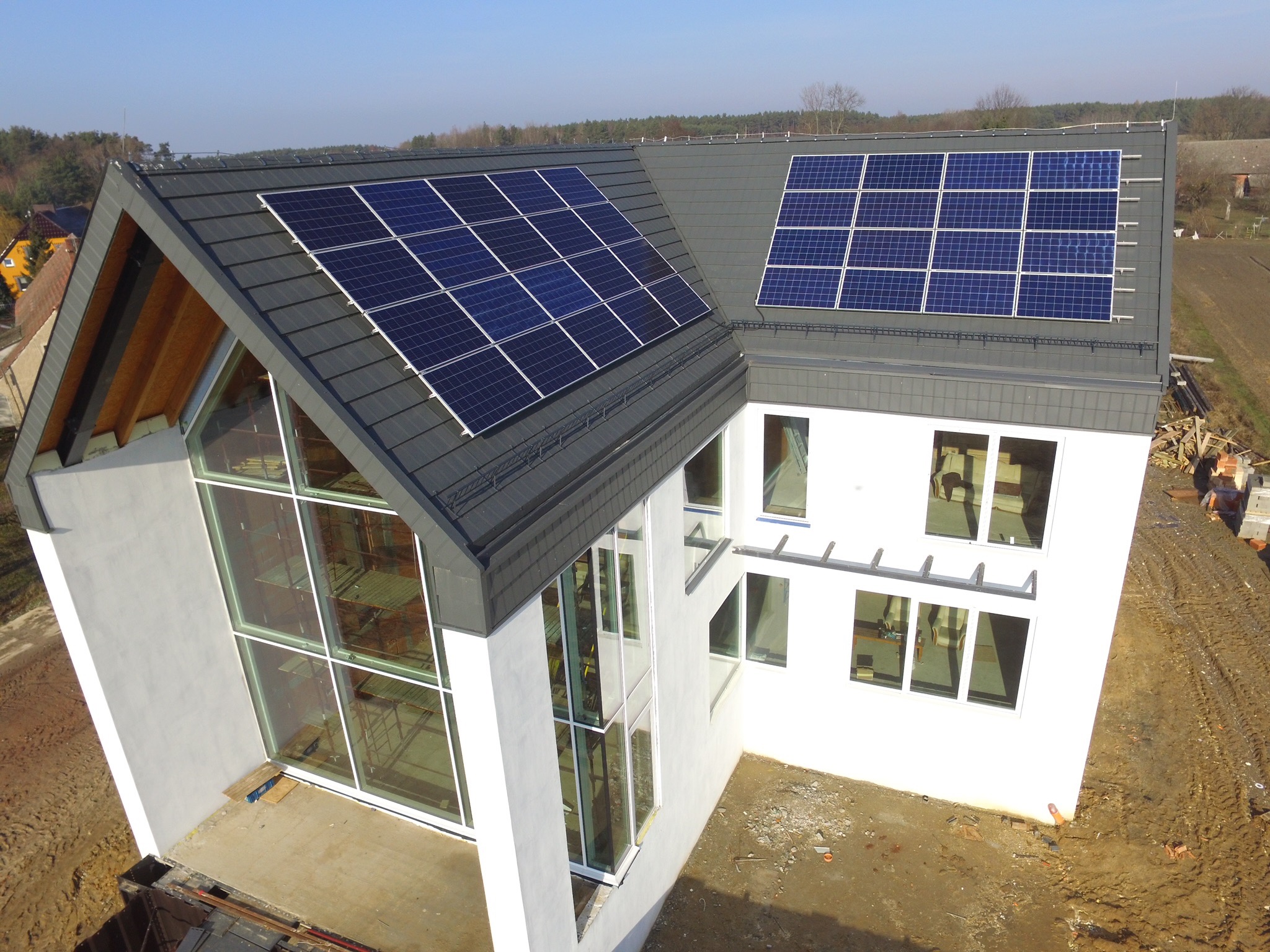 Nowoczesny dom z szarym dachem pokrytym panelami słonecznymi, duże okna z białymi ramami, teren wokół domu w trakcie prac budowlanych.