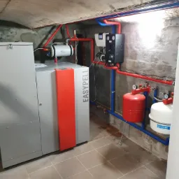 Kotłownia z zainstalowanym piecem na pellet EasyPell, zbiornikami wyrównawczymi i instalacją grzewczą z rur w izolacji, widok na betonowy sufit piwnicy.