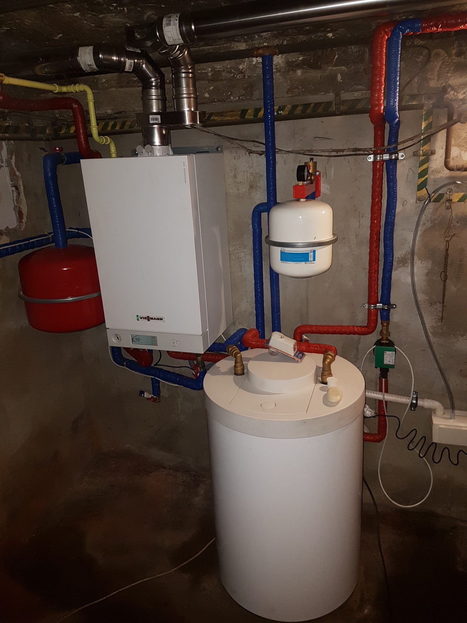 Kocioł grzewczy Viessmann z zasobnikiem ciepłej wody i zbiornikiem wyrównawczym, instalacja z rur w izolacji czerwonej i niebieskiej, widoczne elementy instalacji gazowej i hydraulicznej w piwnicy.