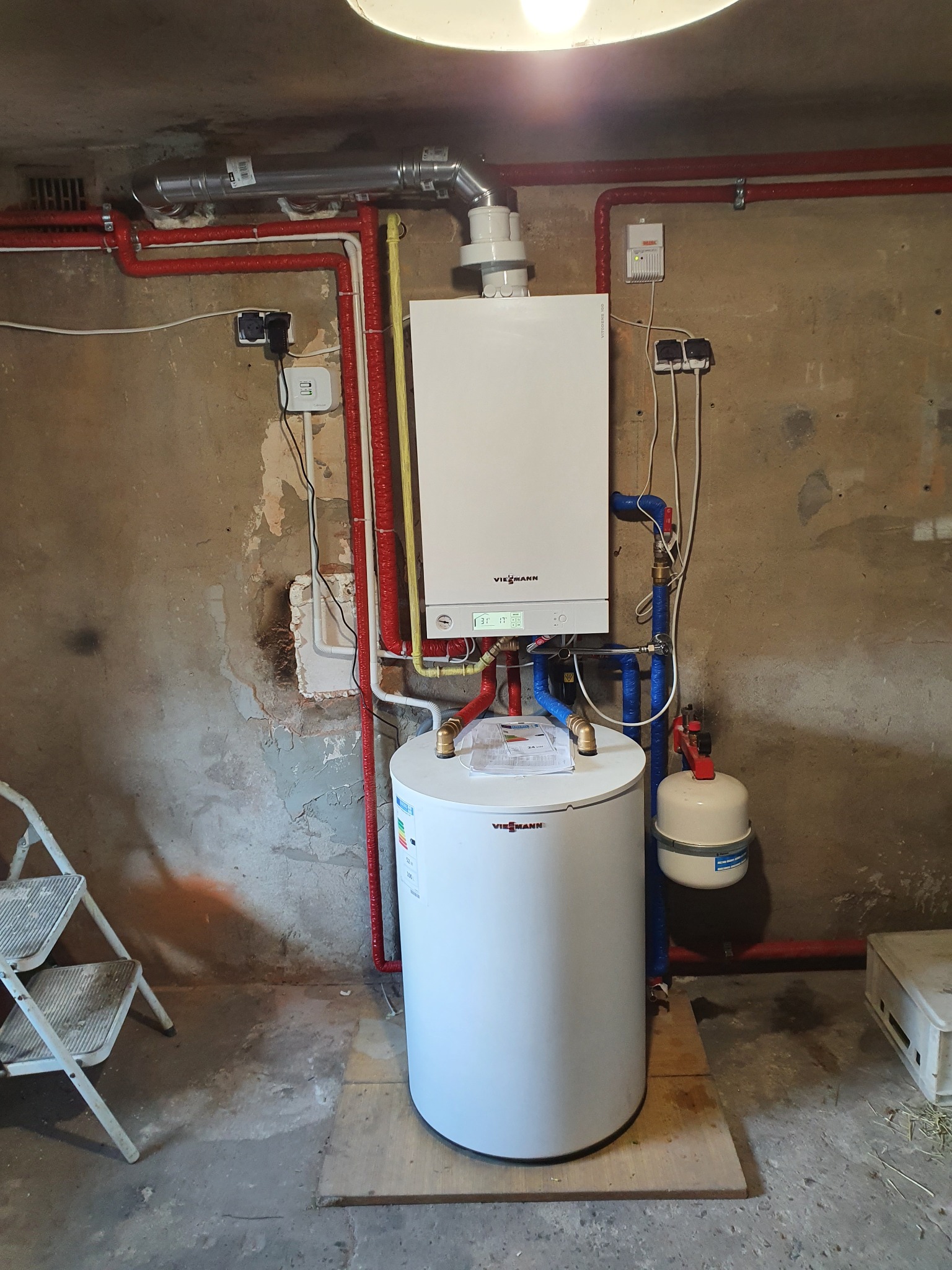 Zainstalowany, wiszący, biały piec gazowy Viessmann z podłączonym zbiornikiem ciepłej wody, rurami grzewczymi w czerwonej izolacji i elementami instalacji w piwnicy o surowych ścianach.