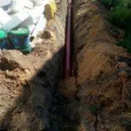 Rów z ułożoną rurą kanalizacyjną z tworzywa sztucznego, widok z góry na wykop w gruncie, w tle kostka brukowa i trawnik.