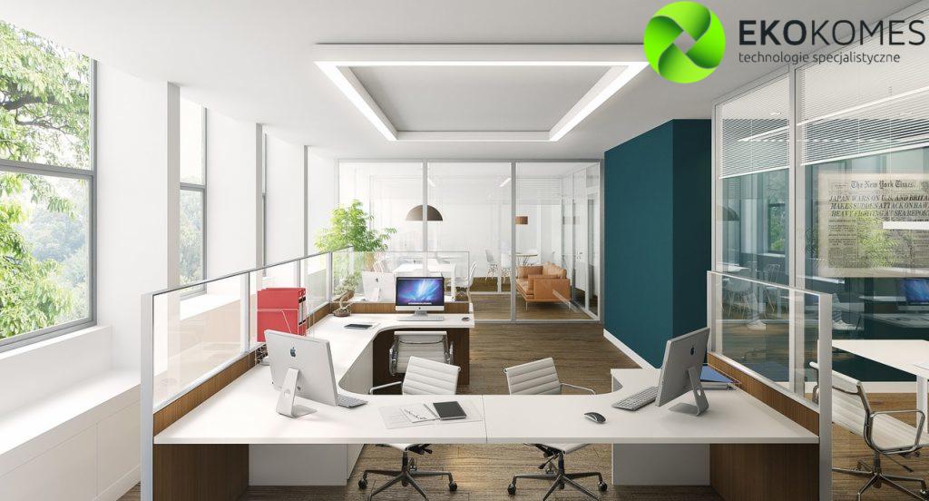 Jasne, nowoczesne biuro z białymi biurkami, komputerami iMac, szklanymi ścianami działowymi i widokiem na zieleń za oknem. Na ścianie wisi oprawiona gazeta The New York Times. W tle widoczna część...