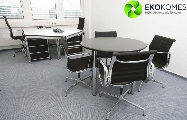 Jasne biuro z szarym dywanem, okrągłym stołem konferencyjnym otoczonym przez cztery krzesła z chromowanymi nogami oraz biurkiem z telefonem, w prawym górnym rogu widoczne logo firmy Ekokomes.