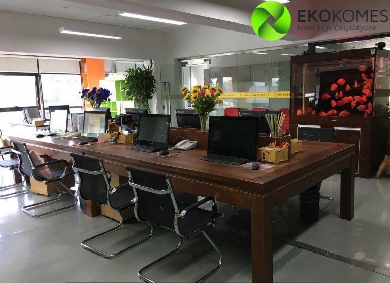 Przestronne biuro z długim drewnianym stołem, krzesłami biurowymi, komputerami, kwiatami i akwarium z czerwonymi rybkami w tle, widoczne logo firmy EKOKOMES.