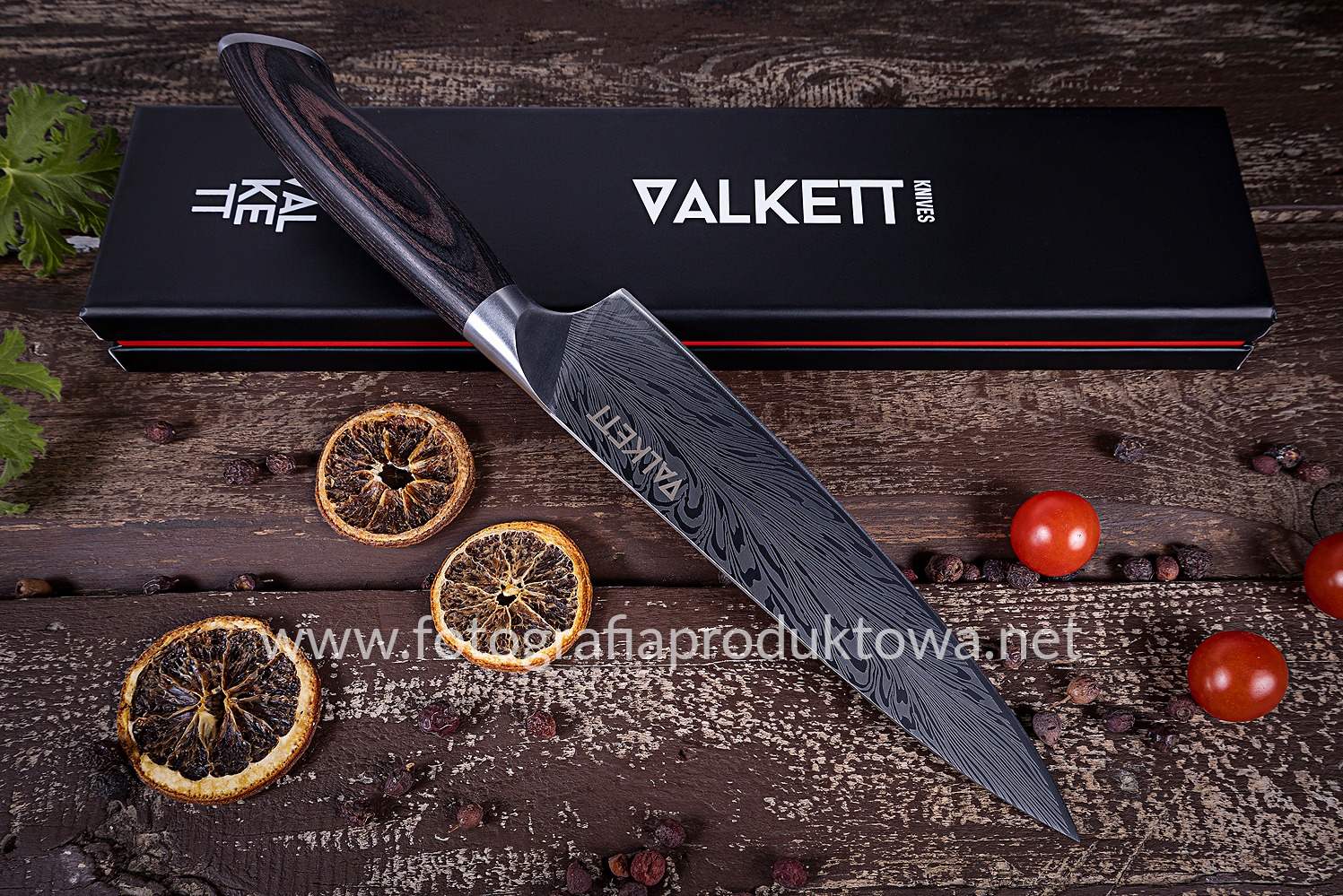 Nóż VALKETTI z wzorem damasceńskim, leżący na rustykalnym drewnianym stole obok suszonych pomarańczy i pomidorków cherry, z pudełkiem w tle. Fotografia produktowa.