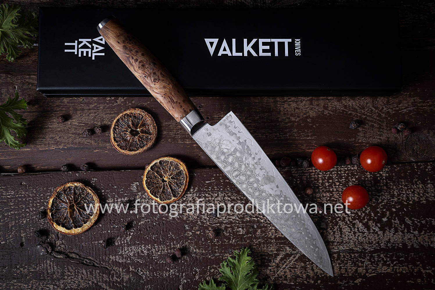 Nóż Valkett z wzorem damasceńskim leży na drewnianym stole z suszonymi pomarańczami i pomidorami, obok czarne pudełko z logo firmy. Fotografia produktowa.