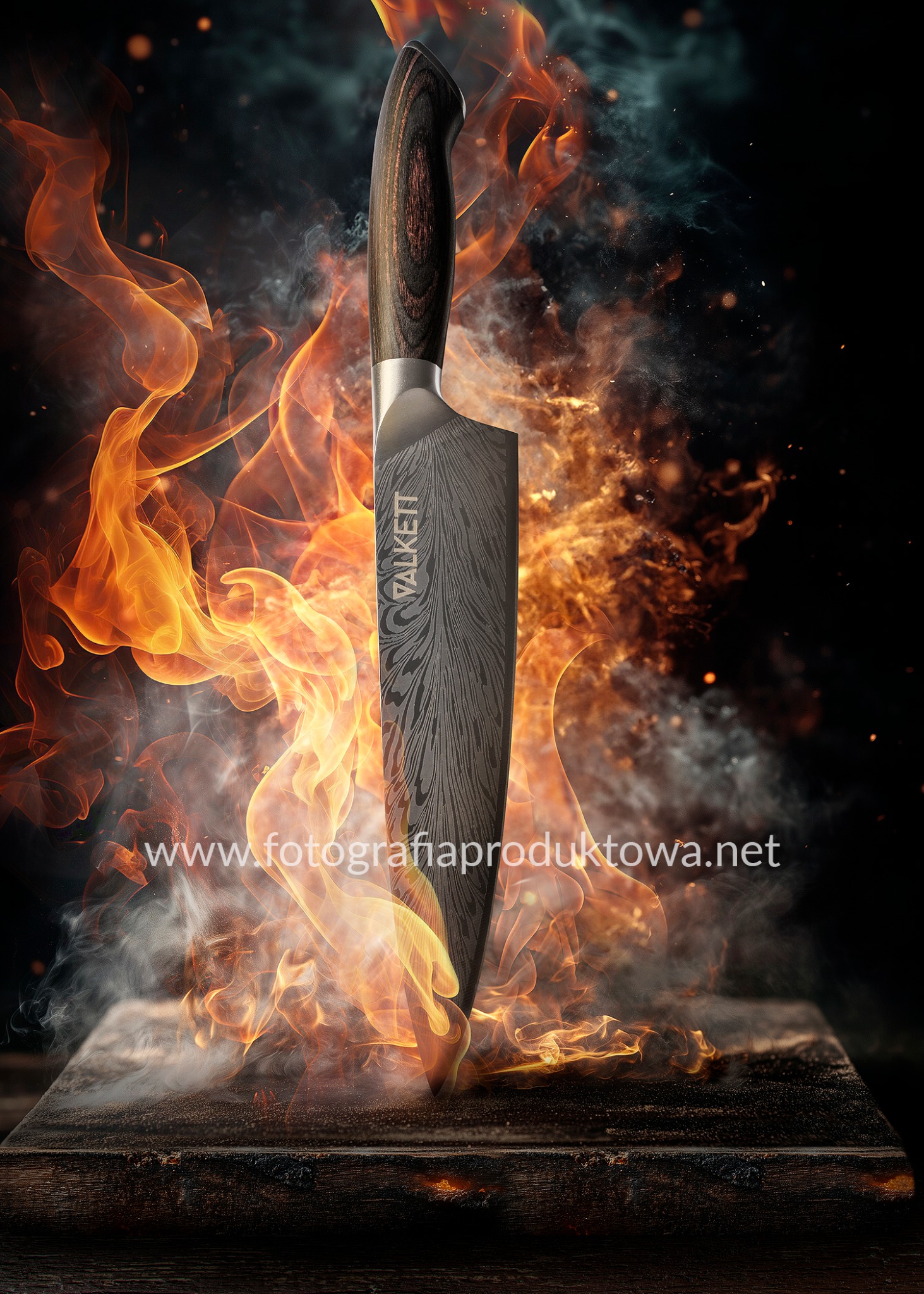 Nóż Valkett wbity w drewniany blok, otoczony płomieniami i dymem. Ostrze z widocznym wzorem damasceńskim. Ujęcie z niskiej perspektywy.