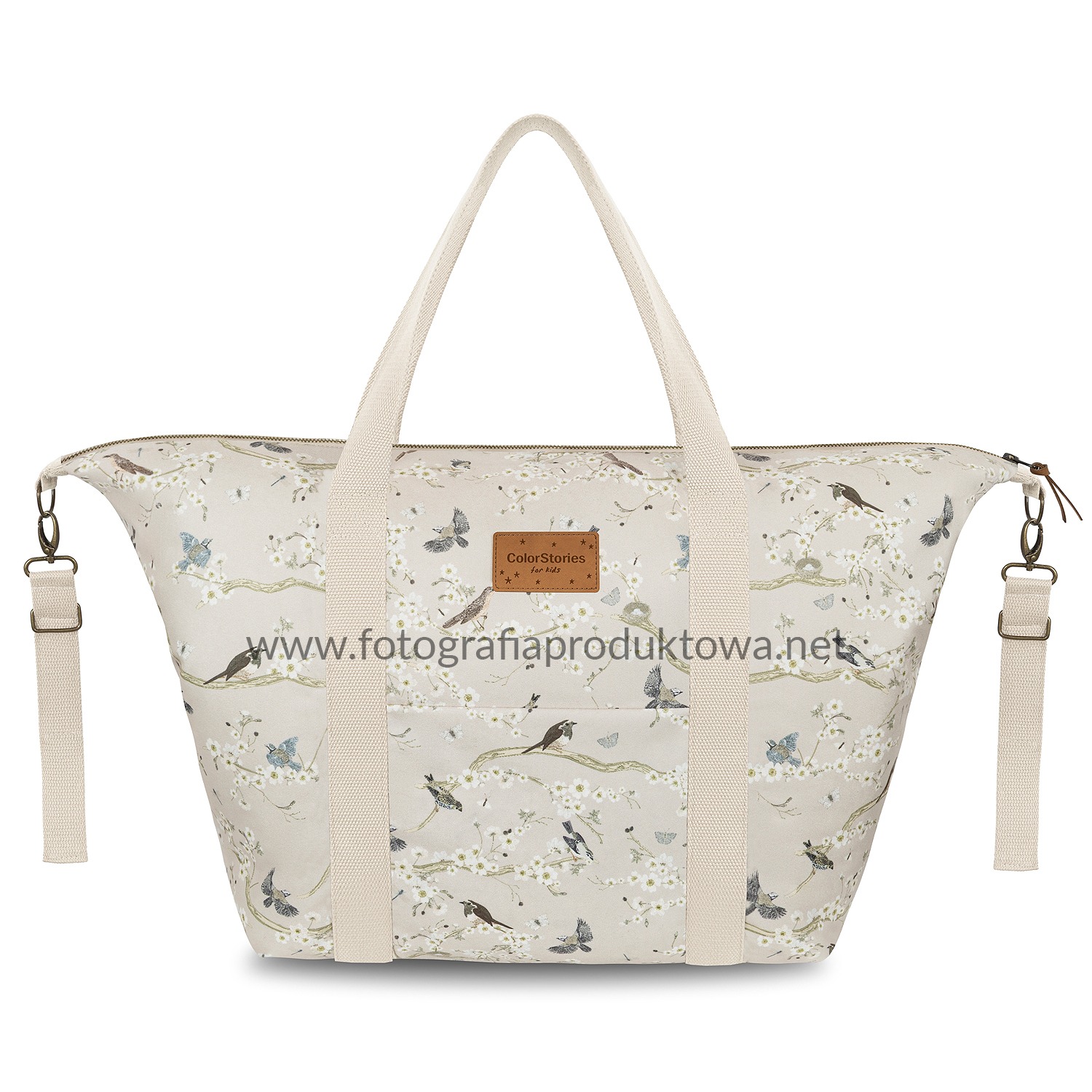 Beżowa torba podróżna z motywem ptaków i kwiatów, z jasnymi uchwytami, na białym tle. Subtelne oświetlenie podkreśla wzór. Widoczny zamek i elementy regulacji.