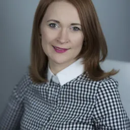 Portret uśmiechniętej kobiety z rudymi włosami w czarno-białej koszuli w kratę z białym kołnierzykiem, na jednolitym szarym tle.