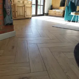 Ułożona w jodełkę podłoga z paneli drewnopodobnych, widok na salon z drewnianymi meblami i turkusowym fotelem.