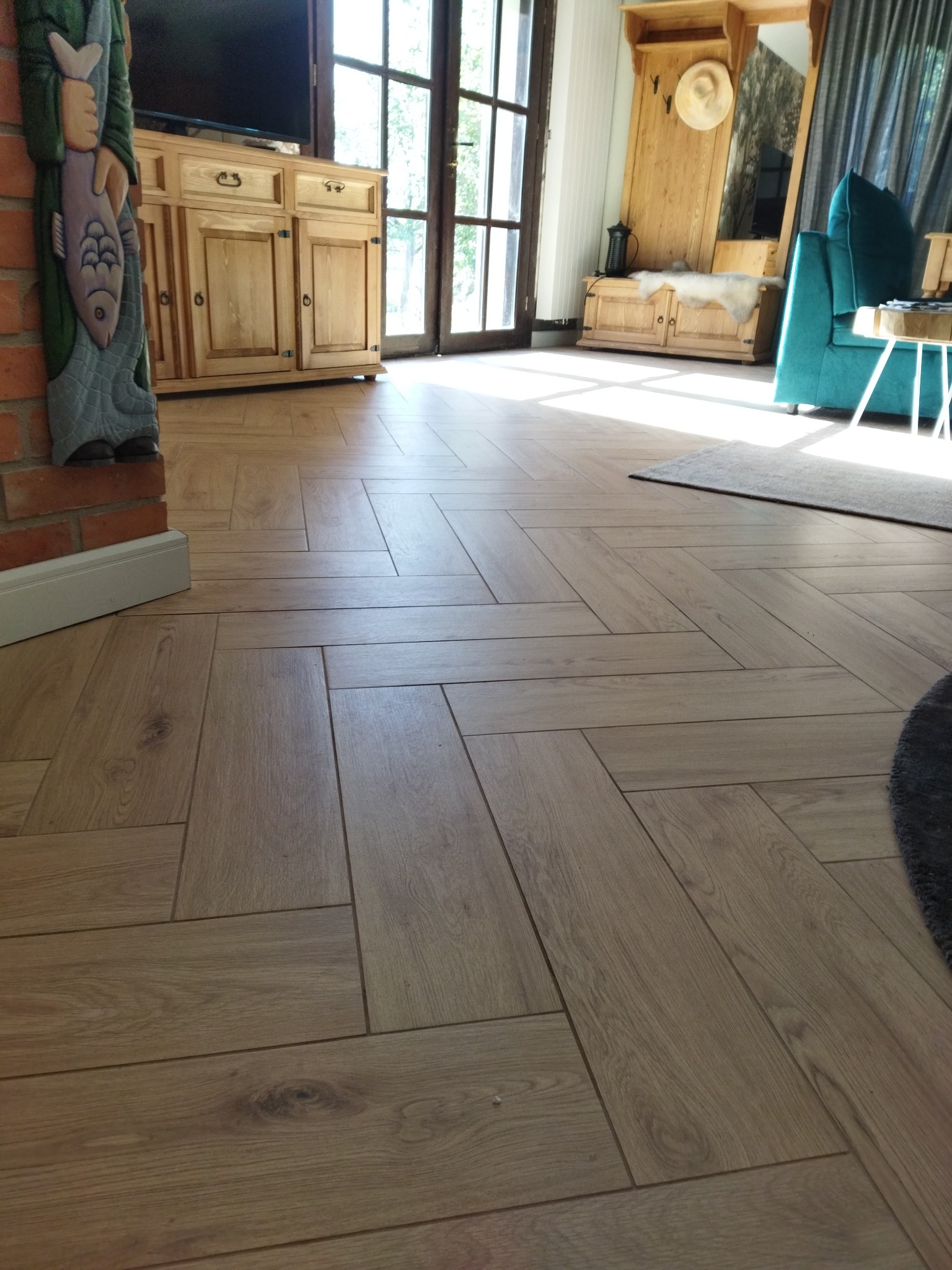 Ułożona w jodełkę podłoga z paneli drewnopodobnych, widok na salon z drewnianymi meblami i turkusowym fotelem.