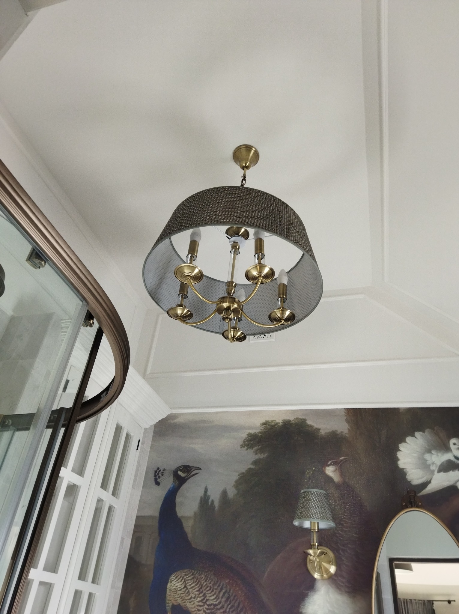 Elegancki żyrandol z abażurem w odcieniach szarości i złotymi elementami, widoczny na tle białego sufitu z dekoracyjnymi listwami oraz fragmentu ściany z malowidłem przedstawiającym pawia.