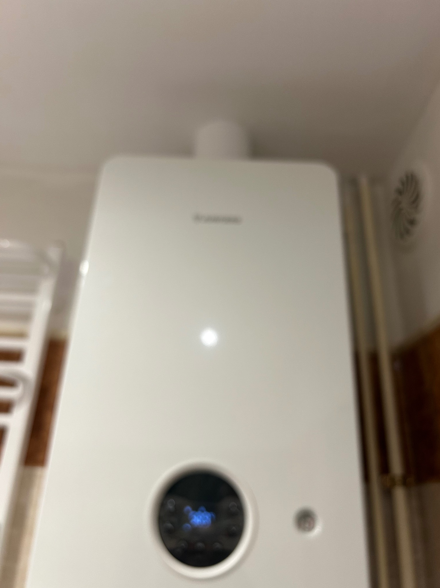 Biały, wiszący piec gazowy z wyświetlaczem, zamontowany na ścianie w pomieszczeniu. Widoczny fragment grzejnika drabinkowego i kratka wentylacyjna.