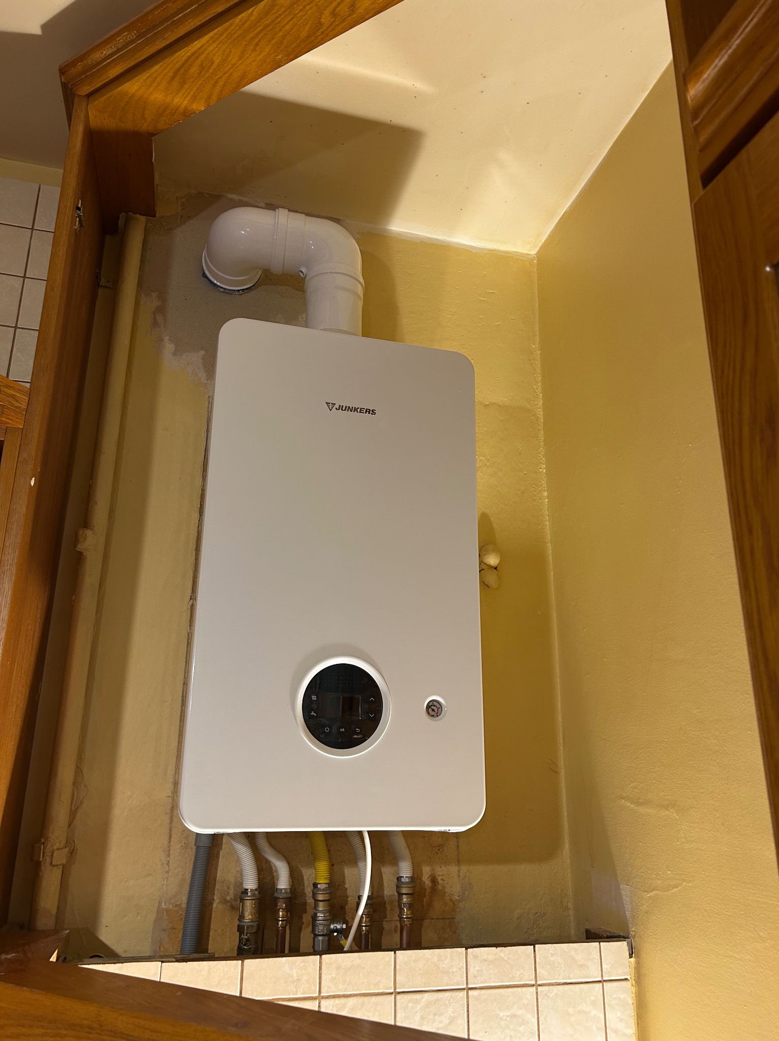 Biały, nowoczesny piec gazowy Junkers zamontowany na żółtej ścianie w rogu pomieszczenia, widoczne rury doprowadzające gaz i wodę, oraz fragment drewnianej zabudowy.