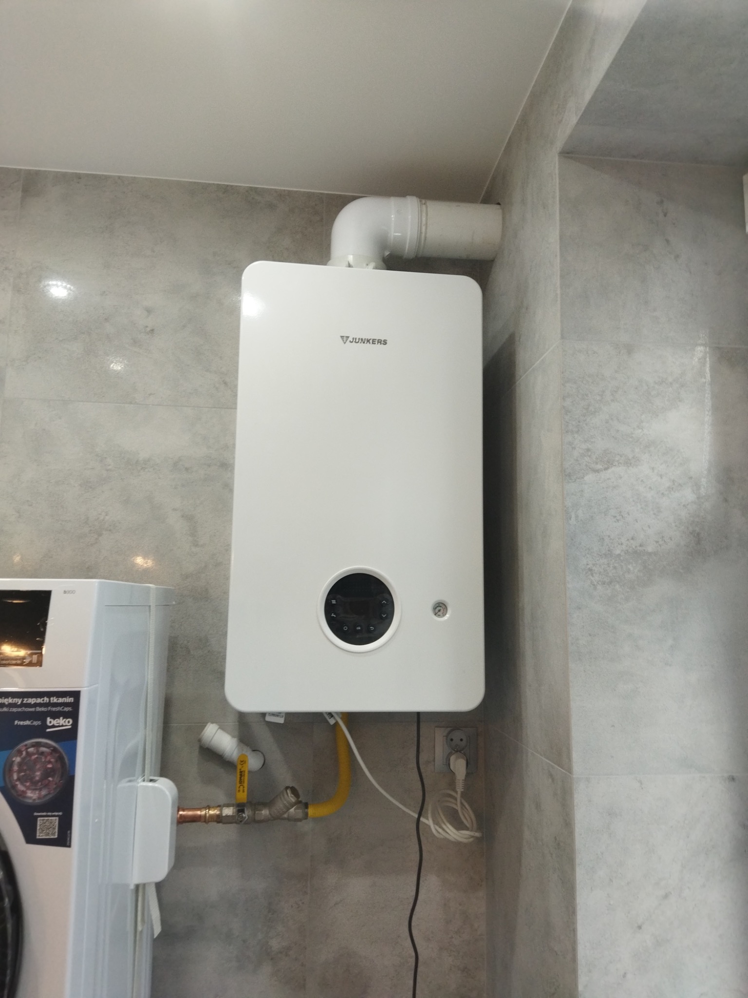 Biały, wiszący piec gazowy Junkers na szarej, płytkowej ścianie w łazience, z widocznymi przyłączami gazowymi i elektrycznymi oraz fragmentem pralki Beko.
