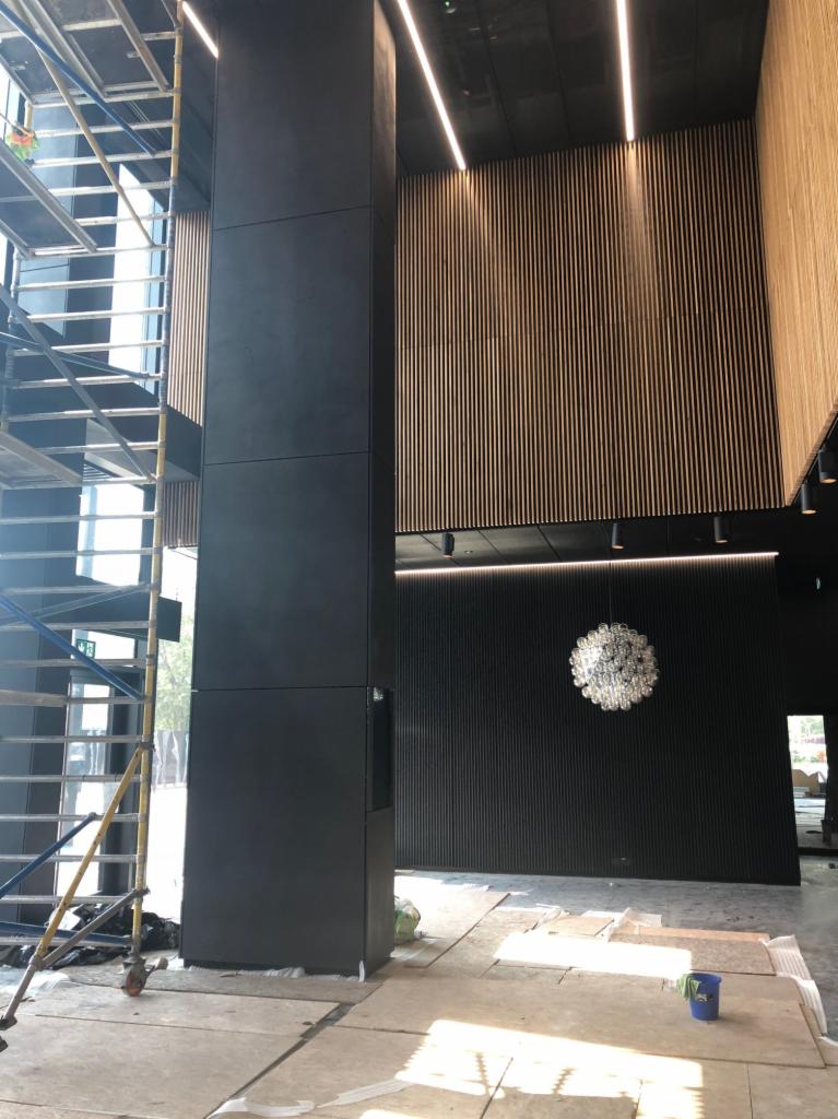 Wnętrze budynku KTW w trakcie wykańczania: widoczne rusztowania, kolumna obłożona płytami, drewniana boazeria na ścianach i sufitach, oraz designerski żyrandol.