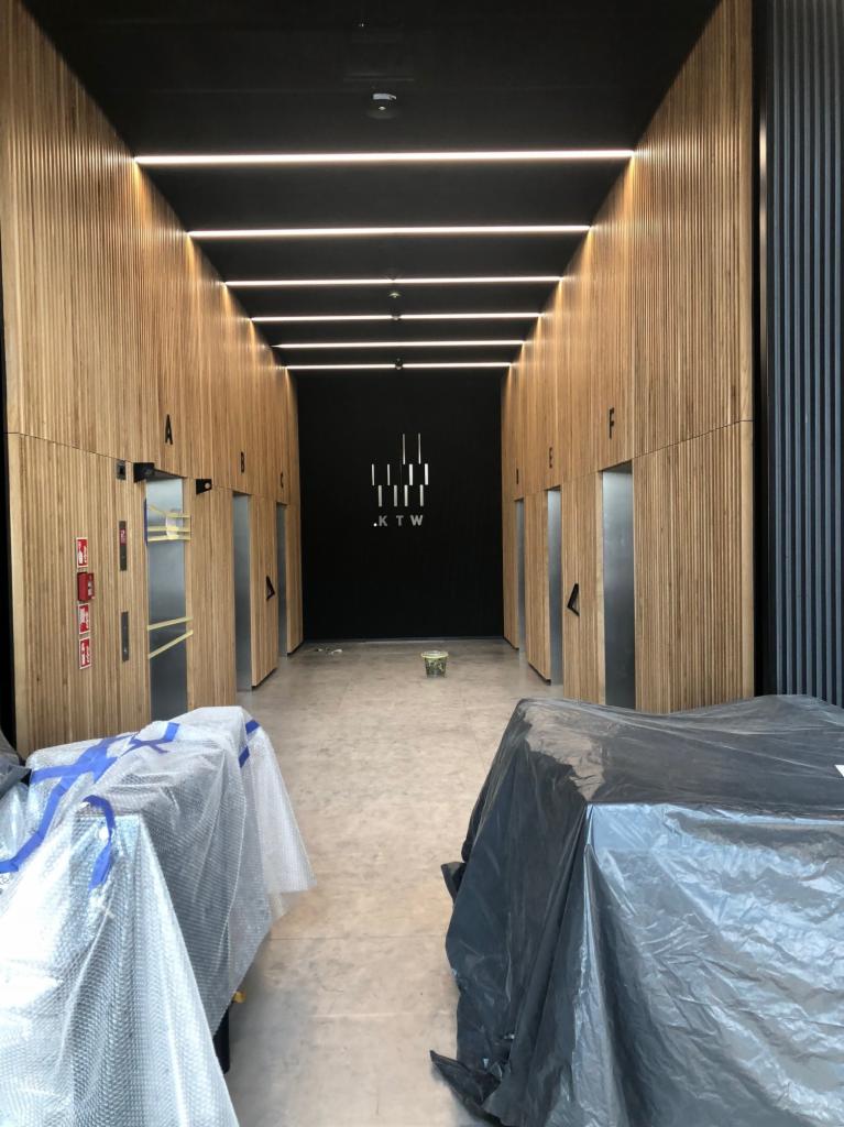 Wnętrze budynku KTW w trakcie otwierania: korytarz z drewnianymi panelami, windami i czarną ścianą z logo, zabezpieczone meble folią ochronną.