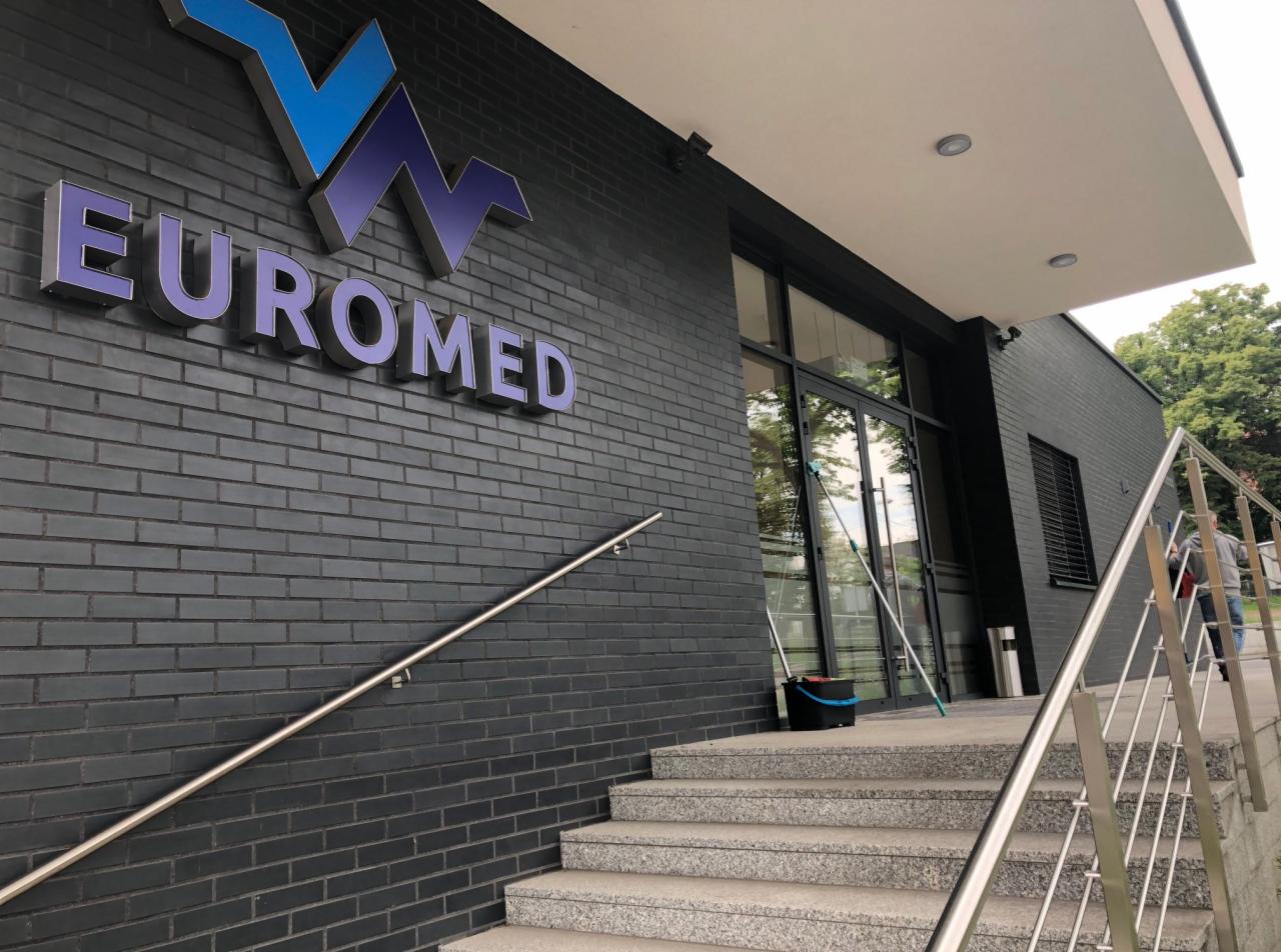 Budynek Euromed z ciemnej cegły z logo, widoczne schody wejściowe, poręcze i sprzęt do mycia okien oparty o szybę.