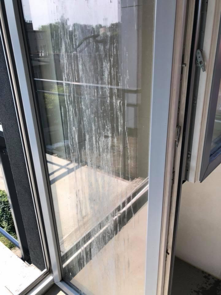 Bardzo brudne okno balkonowe z zaciekami po deszczu i osadami, widok z zewnątrz na zieloną roślinność.