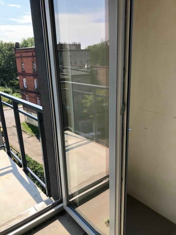 Widok z balkonu przez otwarte, przesuwne okno; w odbiciu widać budynek z czerwonej cegły i zieleń; balkon betonowy.