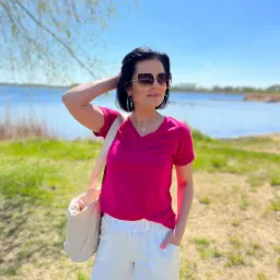 Kobieta w malinowej koszulce i białych spodniach pozuje na tle jeziora w słoneczny dzień, prezentując casualowy strój.