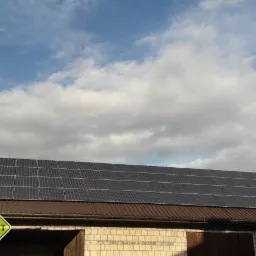 Panele słoneczne na brązowym dachu budynku gospodarczego z białej cegły, na tle pochmurnego nieba. W lewym dolnym rogu logo firmy Segarit.