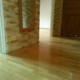 Wypolerowana drewniana podłoga z widocznymi deskami parkietu, ściana wyłożona imitacją kamienia, fragment drewnianych drzwi w tle.