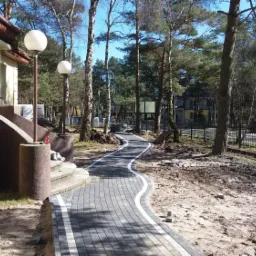 Ścieżka z szarej kostki brukowej z białym obramowaniem, wijąca się między drzewami i budynkiem w słoneczny dzień, widoczne niedokończone prace i stos kostki przy schodach.