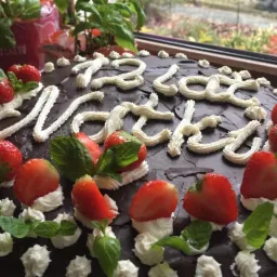 Ciemny tort z kremowym napisem i dekoracją z truskawek oraz listków mięty, widok z góry przy oknie.