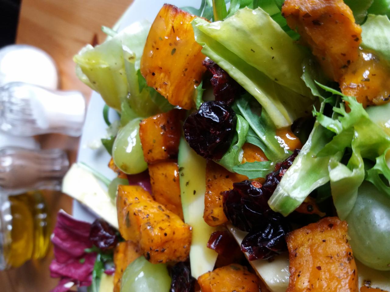 Apetyczna sałatka z pieczonej dyni, zielonych winogron, żurawiny i mieszanki sałat, polana sosem, widok z góry z boku, z przyprawnikiem w tle.