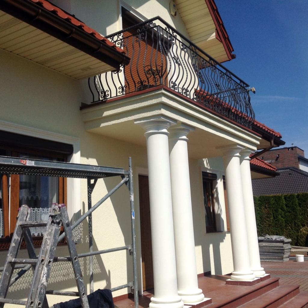 Elewacja domu jednorodzinnego z kolumnami i balkonem z kutą balustradą, widoczny fragment rusztowania, świeżo pomalowana elewacja w jasnym odcieniu.