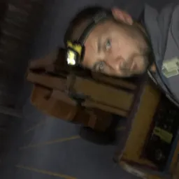 Elektryk z czołówką oparty o skrzynkę narzędziową w ciemnym pomieszczeniu. Widoczny fragment instalacji elektrycznej w tle. Niefortunny kadr.