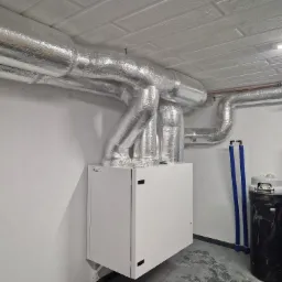 Biała centrala wentylacyjna z izolowanymi przewodami wentylacyjnymi w pomieszczeniu technicznym, obok zbiornik ciśnieniowy i piec Vaillant z miedzianą instalacją.