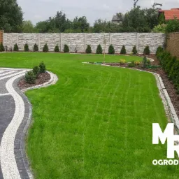 Zadbany trawnik z dekoracyjnym obrzeżem z kostki granitowej, ozdobne nasadzenia z tuj i krzewów, płot betonowy i drewniany w tle.