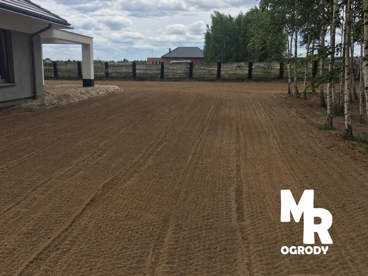 Przygotowany teren pod trawnik, świeżo wyrównana ziemia z widocznymi śladami po maszynach, obok domu z białym podcieniem i rzędem brzóz wzdłuż ogrodzenia.