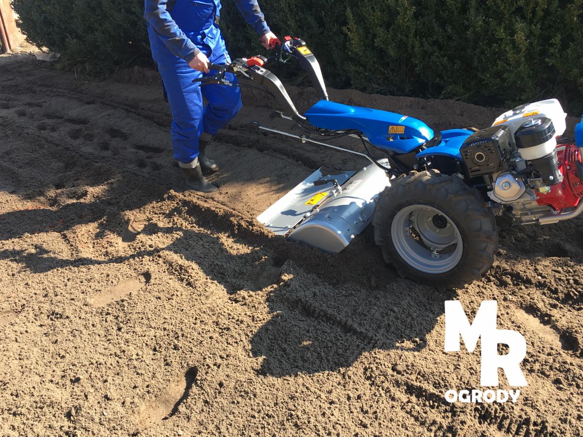 Operator w niebieskim kombinezonie obsługuje glebogryzarkę spalinową, przygotowując ziemię pod trawnik; widoczne ślady kół i stóp w świeżo spulchnionej glebie.