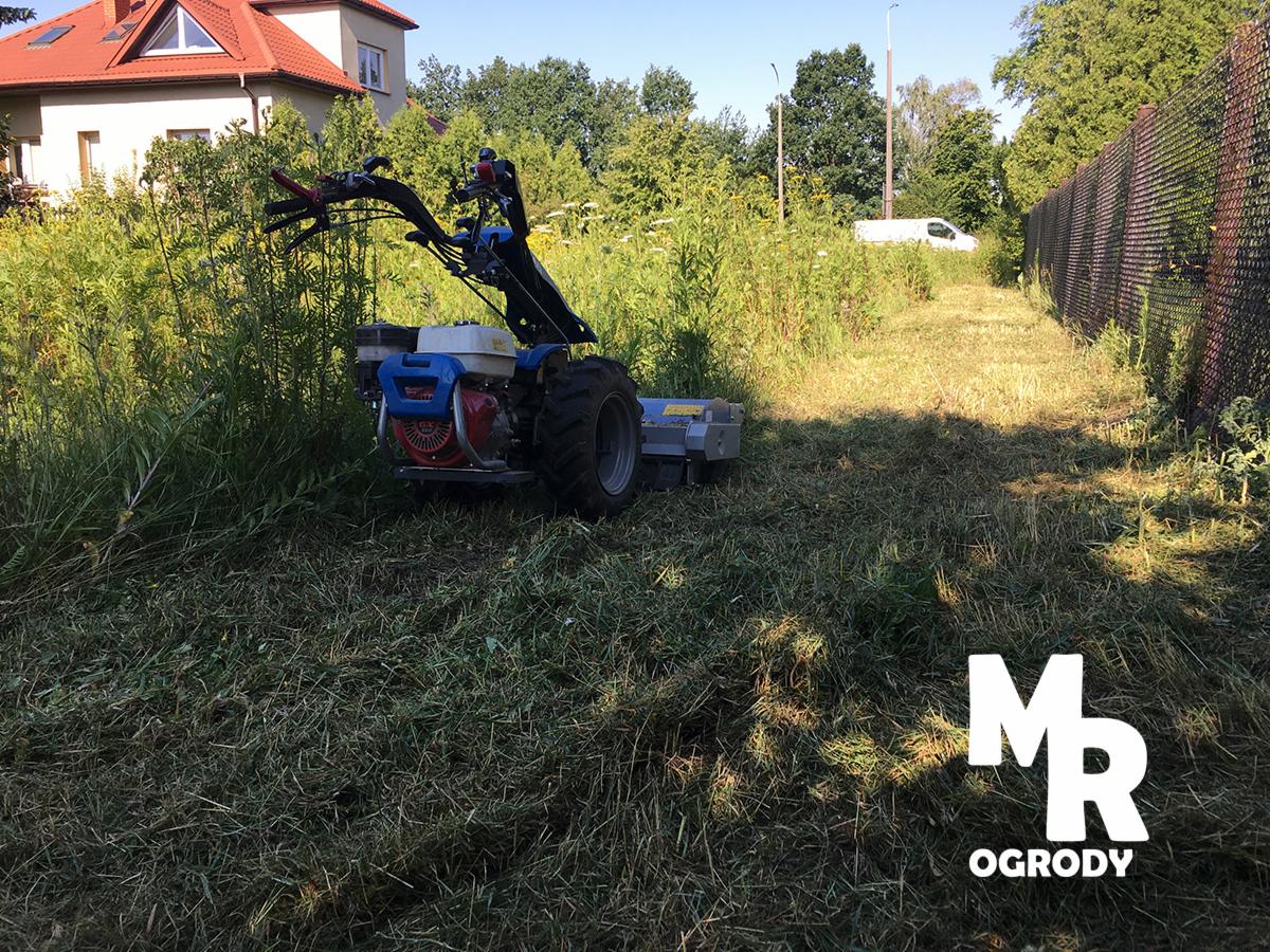 Profesjonalna kosiarka rotacyjna w trakcie pracy na zarośniętym terenie przy płocie, z widocznym budynkiem mieszkalnym w tle i logo firmy ogrodniczej w prawym dolnym rogu.