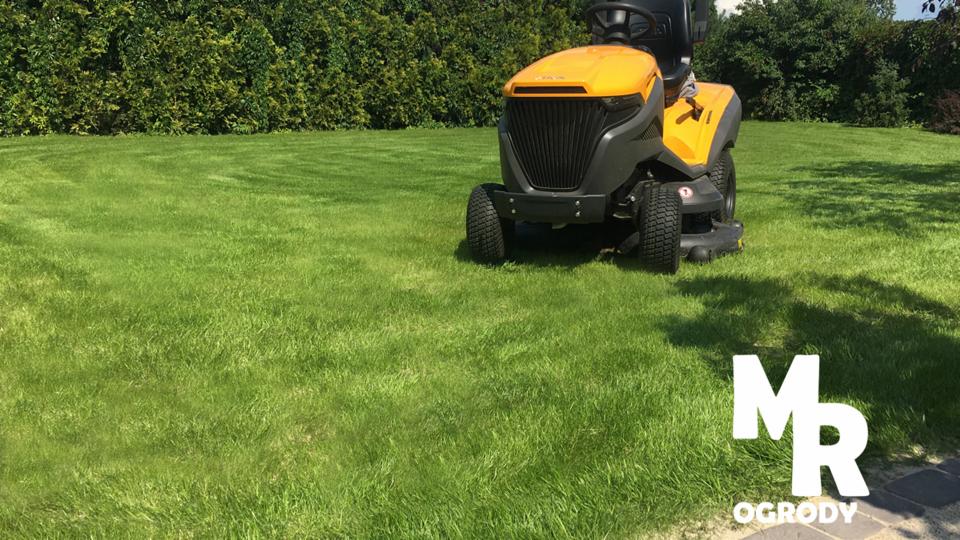 Żółto-szara kosiarka traktorek na idealnie przystrzyżonym trawniku z widocznym wzorem koszenia, żywopłot w tle, logo firmy w prawym dolnym rogu.