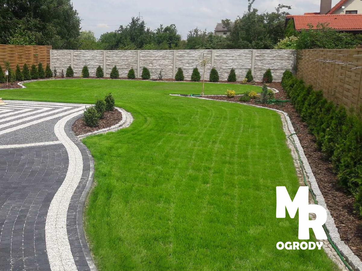 Zadbany trawnik z dekoracyjnym obrzeżem z kostki granitowej, ozdobne nasadzenia z tuj i krzewów, płot betonowy i drewniany w tle.