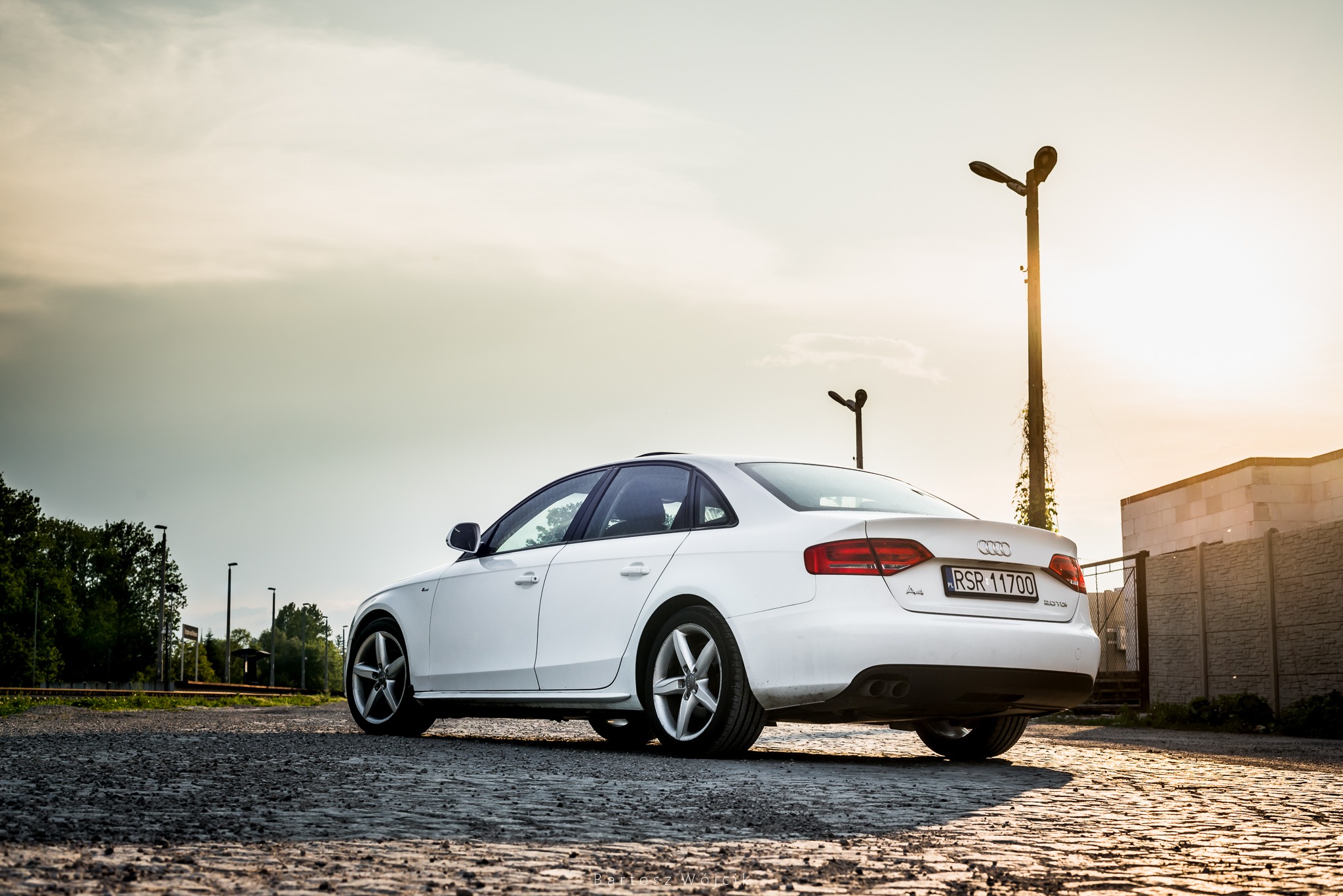 Białe Audi A4 na brukowanej drodze, widok z tyłu, oświetlone zachodzącym słońcem, słupy oświetleniowe w tle.