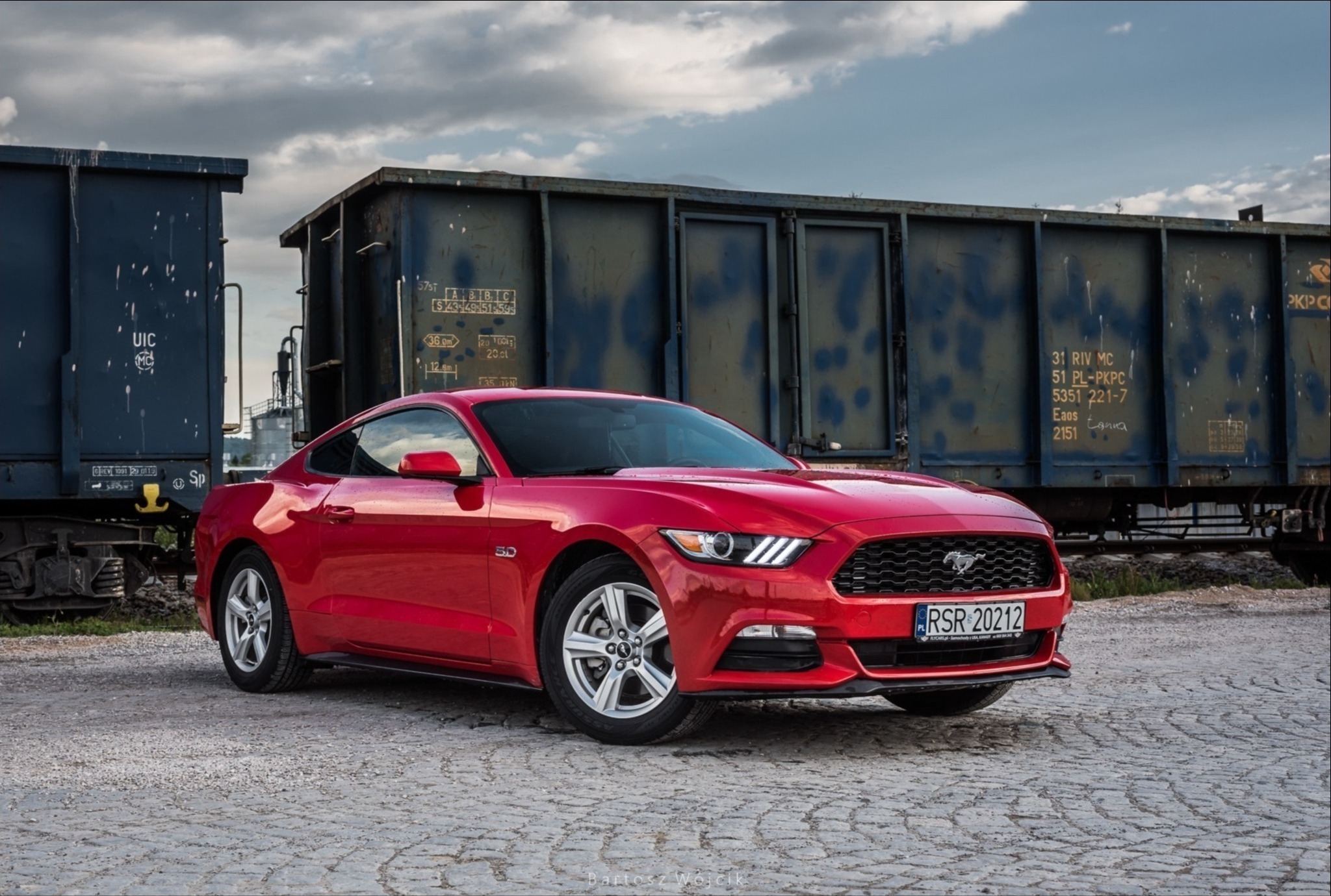 Czerwony Ford Mustang na tle starych wagonów kolejowych, sfotografowany na brukowanej nawierzchni w pochmurny dzień.