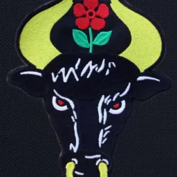 Czarna naszywka z wyhaftowaną głową byka z czerwonymi oczami, żółtymi rogami i kolczykiem w nosie, nad głową czerwony kwiat na zielonej łodydze.