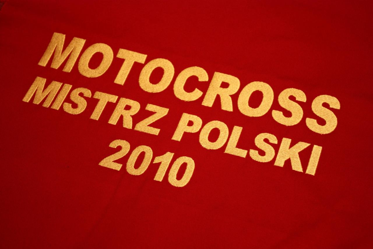 Czerwona koszulka z wyhaftowanym złotym napisem 'MOTOCROSS MISTRZ POLSKI 2010', widoczne detale splotu materiału.