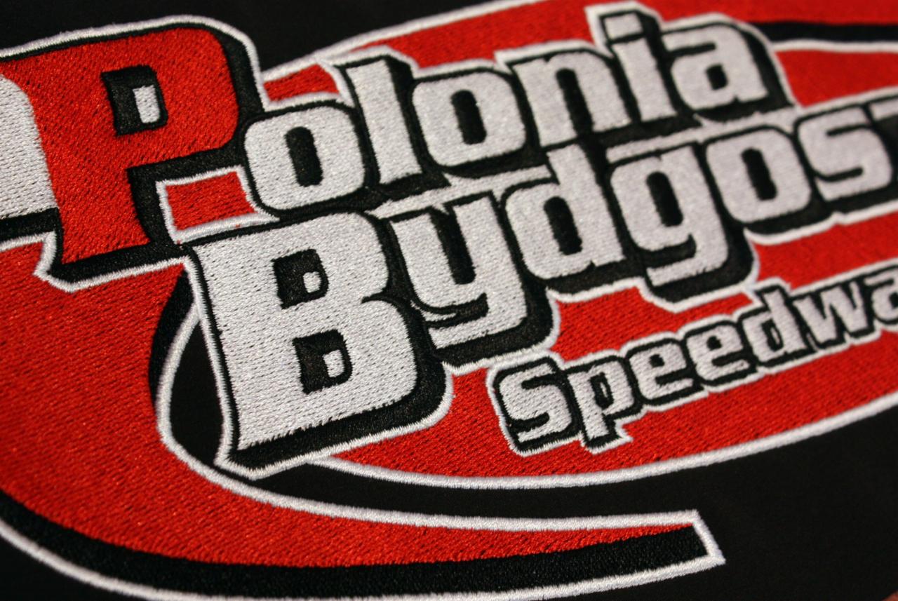 Zbliżenie haftowanego logo 'Polonia Bydgoszcz Speedway' na czarnym materiale, z wyraźną teksturą nici w kolorach czerwonym, białym i czarnym.
